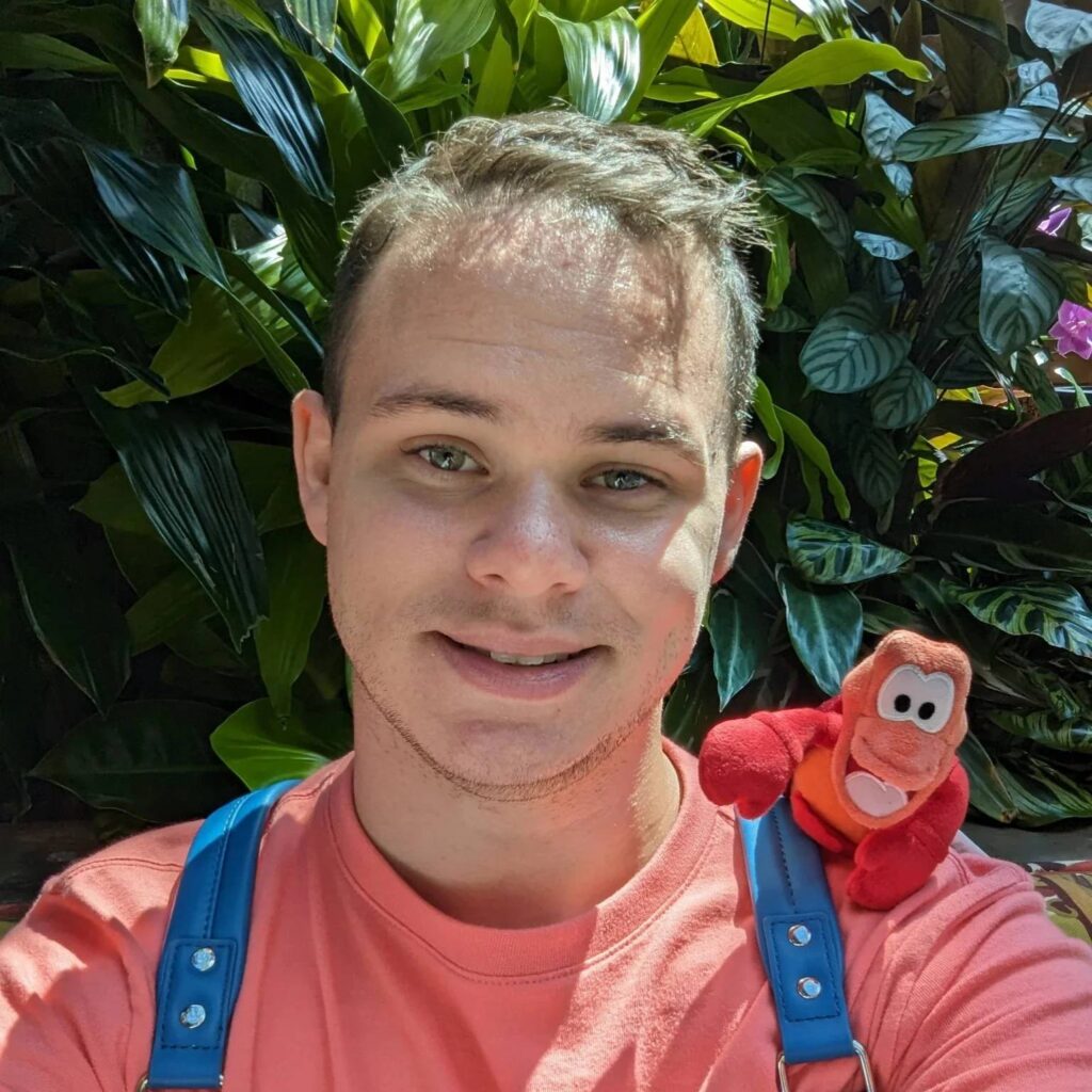 Matthew Manganaro with a stuffed animal of Sebastian on his shoulder standing in front of a plant.